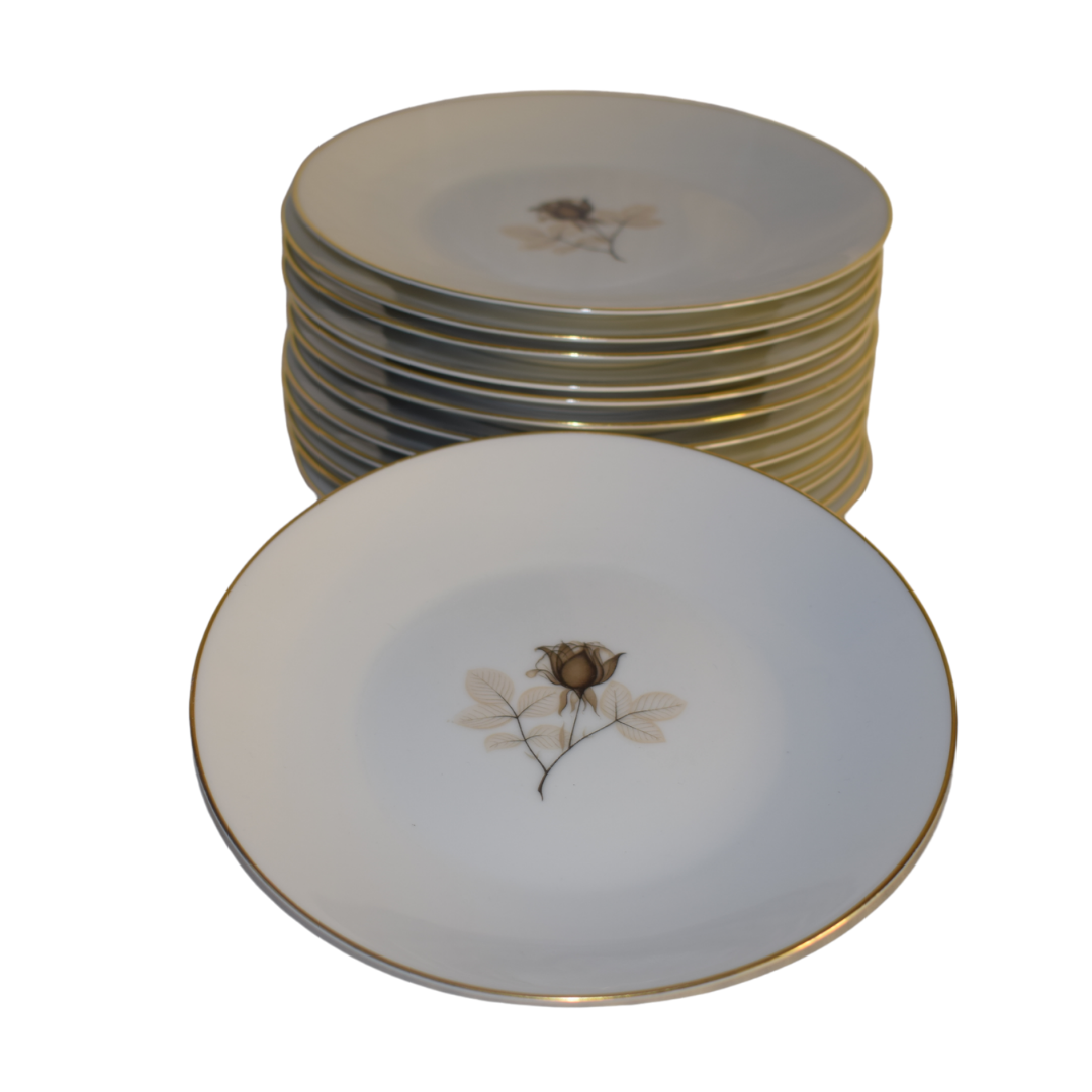 Et sæt hvide porcelænstallerkener med guldkant og et delikat rosemotiv i midten.